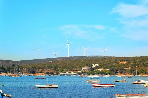 Wind turbines on a hill in the distance overlooking the ocean, filled with boats.